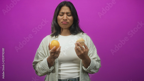 Fotografie Woman holding an orange and a muffin, grimaces while comparing choices in studio; indecision temptation