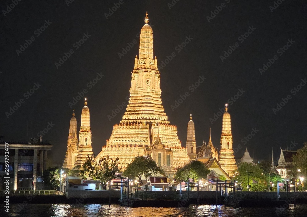 Naklejka premium Illuminated Wat Arun temple with golden spires reflecting on the Chao Phraya River at night in Bangkok, Thailand