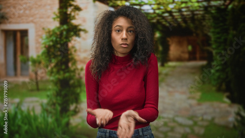 Foto Young woman with curly hair gesturing with bare hands and speaking in front of building courtyard pathway wearing a red turtleneck sweater and jeans; thoughtful reflection