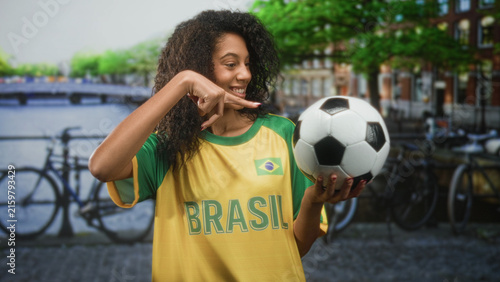 Obraz na plátne Teenage woman with curly hair wearing brasil t shirt holds soccer ball in hand and makes a celebratory fist gesture on street; team pride