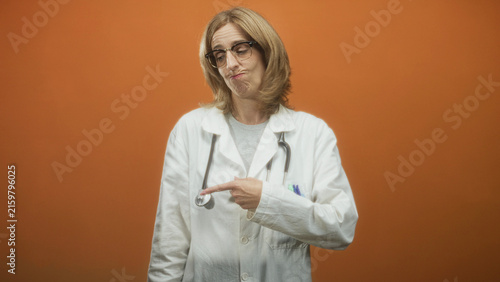 Canvas Print Woman doctor in white lab coat and glasses points finger at stethoscope and pens in chest pocket in studio; wry skepticism