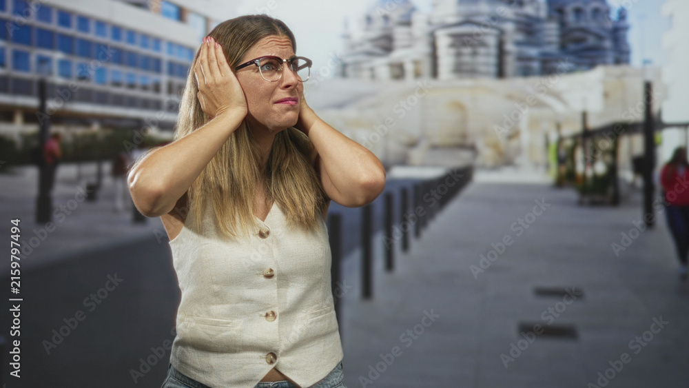 Fototapeta premium Woman covers ears with hands and glasses on street near monument and bollards, wearing sleeveless vest and jeans; stress discomfort.