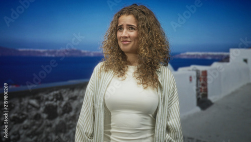 Fotografie Woman wincing with closed eyes showing bust and striped cardigan on a seaside street with whitewashed walls and ocean view; melancholy travel reflection