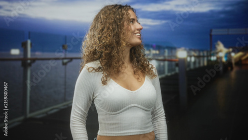 Foto Woman with curly ringlets and visible midriff smiling while looking sideways on a waterfront pier at dusk with glass railings and city lights; serenity