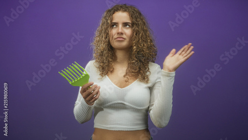 Obraz na płótnie Woman holding a green comb at her waist, right hand raised in a dismissive wave in a studio with purple backdrop and visible curly hair and white crop top; annoyance