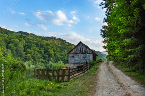 Wooden Barn and Country Road in Mountainous Landscape on Sunny Summer Day