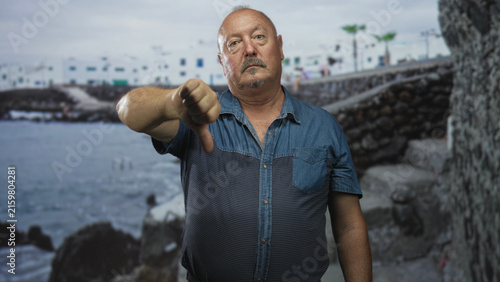 Canvastavla Man with extended hand giving thumbs down gesture by a stone seawall and white building; disapproval rejection