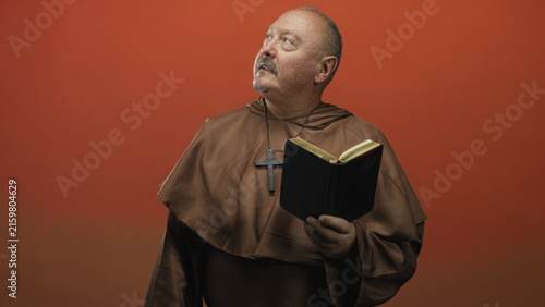 Fotografía Man in brown monk robe holding a book and looking up in a studio with orange backdrop; contemplation prayer serenity