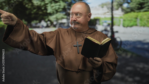 Obraz na plátně Senior hispanic man monk holding bible and cross necklace points finger forward on street path near park; devotion faith guidance