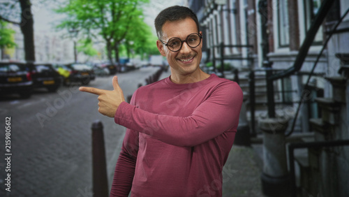 Foto Hispanic man with glasses and moustache points finger to the left on a city street, showing hand and smile; friendly confidence