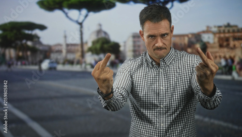Man with moustache and checked shirt shows both middle fingers in street square with stone pavement and umbrella pine trees, confrontational gaze; anger defiance.
