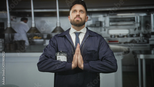 Konstfotografi Man with id badge and tie presses hands together in prayer gesture in a commercial kitchen building; solemn duty