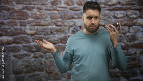 Quadro su tela Man tapping smartphone screen and holding phone to ear in studio against a brick wall; frustration connectivity