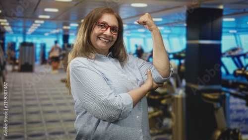 Obraz na płótnie Woman flexing bicep with raised fist and hand holding elbow in gym building, smiling at camera; confidence strength health
