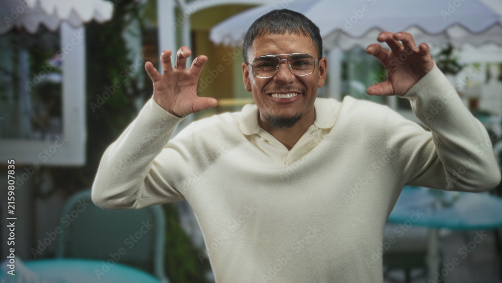 Fototapeta premium Young hispanic man in cream sweater wearing glasses, smiling and raising hands in a playful claw gesture at a street cafe; playful.