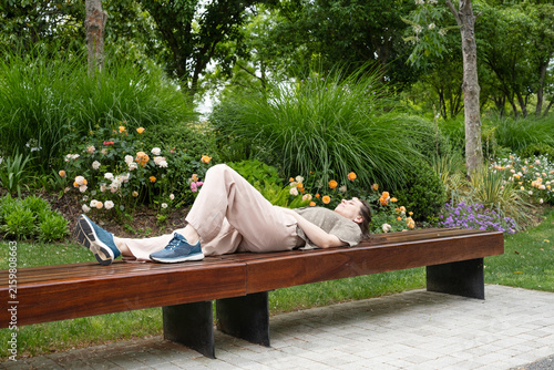 woman lying on bench in blooming park and resting body. relaxed woman enjoying silence among bright flowers and grass. concept of woman leisure and peaceful pause in blooming city garden