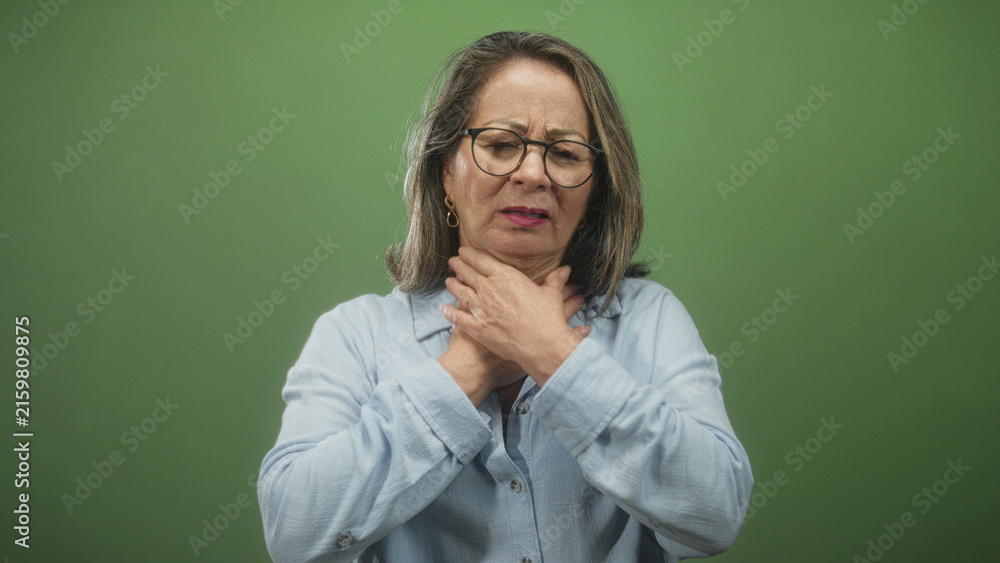Fototapeta premium Hispanic woman with grey hair clutching her throat with both hands in a green studio setting, eyes closed and mouth open showing choking distress; panic medical concern.