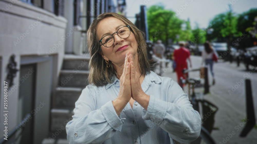 Fototapeta premium Woman with hands pressed palms together in prayer on street beside building stairs, wearing glasses and light blue shirt; serenity.