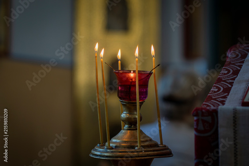 Lit beeswax candles surrounding a red glass vigil lamp in an ornate metal candle stand inside a church, creating a warm and contemplative atmosphere for prayer and worship. 