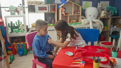 Konstfotografi Girl uses toy stethoscope and syringe on seated boy at a preschool play table in building; playful roleplay learning