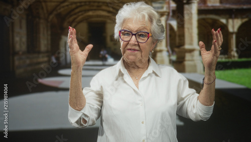 Obraz na plátně Elderly woman raises hands with open palms and smiles in a cloister courtyard beneath stone arches; storytelling warmth