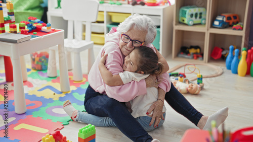 Принти на полотні Elderly woman reaches arms to hug little girl on a toy strewn playmat inside a building; affection bonding
