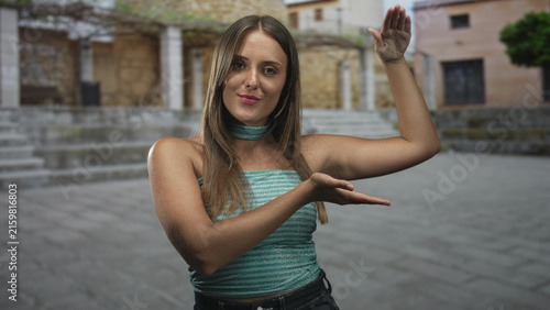 Canvastavla Young brunette woman with bare arms presenting palms as if holding an invisible object, posing in a stone plaza in front of a building; curiosity