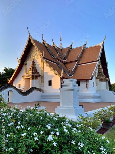 Fotografie Scenic view of the unique cruciform ordination hall at Wat Phumin under blue sky, surrounded by white flowers, landmark of Nan province