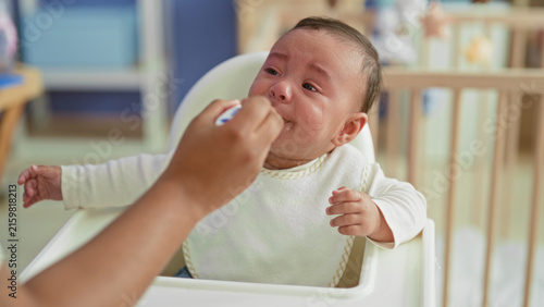 Foto Baby boy crying and spitting spoonful of puree into mouth with messy bib while seated in highchair in building; feeding frustration