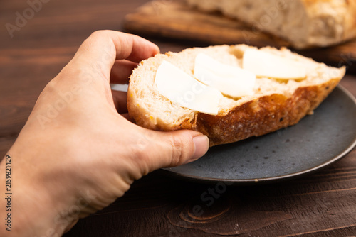 Slice of homemade bread with butter on plate with hand on brown wooden, close up, side view, selective focus.