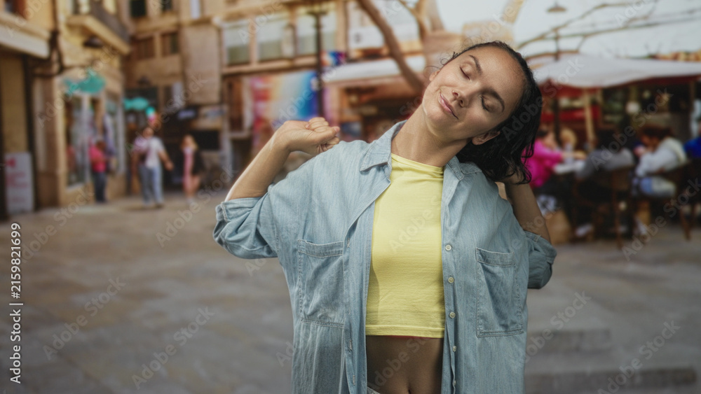 Fototapeta premium Woman pulls denim shirt collar and stretches shoulders with eyes closed in a busy street cafe; relaxed vacation.