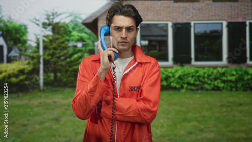 Bild auf Leinwand Man prisoner in orange jumpsuit holds blue handset to ear outside building, number visible on chest; isolation tension
