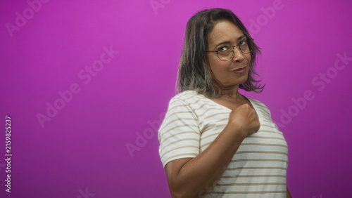 Принти на полотні Hispanic middle age woman points finger to chest, wearing striped shirt and glasses against a magenta studio backdrop and soft lighting; confidence affirmation