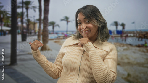 Woman in beige sweater finger to lips for silence and thumb to chest on a street; playful hush mischief.