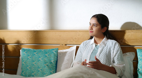 Indian young adult gen z relax calm woman sitting on cozy bed indoor room home place. Girl hold cup mug drink hot tea coffee spend break time do self care enjoy day take job rest