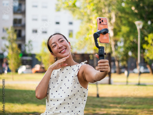 Smiling content creator filming herself with a smartphone stabilizer during a sunny day in the park.