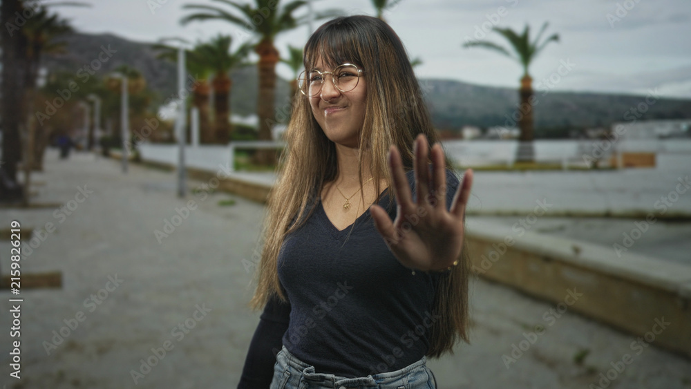 Fototapeta premium Young woman wearing glasses with outstretched hand making a stop gesture toward camera on a street promenade lined with palm trees and benches; playful refusal.
