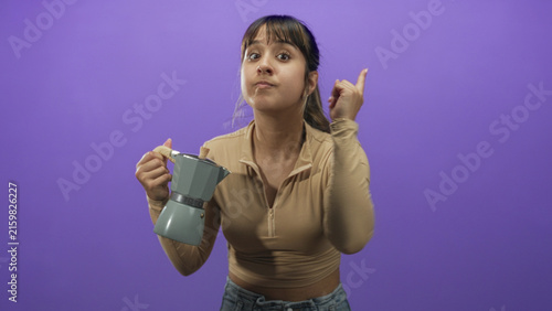 фотографія Young hispanic woman holding gray moka pot with wooden handle points finger to camera in studio set; demanding attention