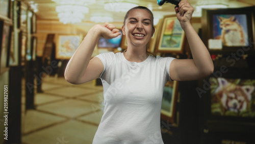 Фотография Woman holding gamepad raises arms and laughs in an art gallery building with framed paintings and chandeliers visible; victory joy