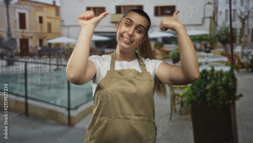 Woman in apron points thumb to herself and makes lively hand gestures in a busy street cafe setting; friendly confidence.