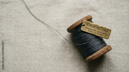 A vintage spool of dark grey thread unraveling on a textured linen surface with an inspirational tag.