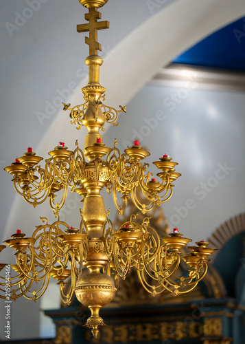 Ornate gilded polyeleos chandelier with numerous candle holders and red candles inside an Orthodox church, featuring intricate metalwork and a prominent cross. Golden light and intricate craftsmanship