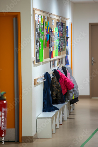 Un couloir d'école maternelle avec un espace de vestiaire, des patères fixées au mur, des bancs de rangement blancs et des panneaux d'affichage d'activités d'enfants.