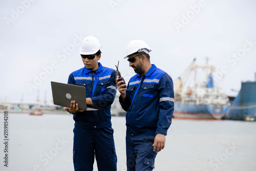 Two industrial workers in protective gear using a laptop and radio at a shipyard, coordinating operations beside cargo vessels and cranes on a busy seaport waterfront.