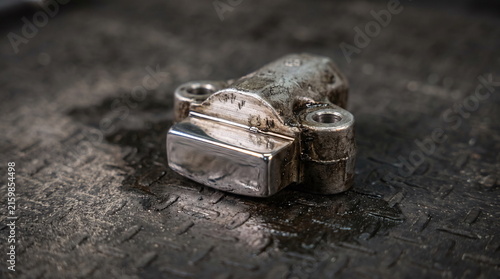 Used metallic automotive timing chain tensioner resting on an oily dark metal diamond plate surface