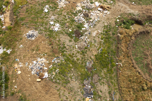 High angle aerial view of an illegal wasteland dump site with scattered plastic garbage and construction debris polluting the natural green field under sunlight.