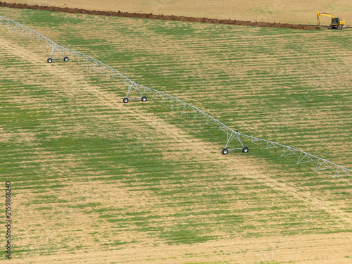 High angle aerial view of modern automated irrigation system and yellow excavator on agricultural farmland under bright daytime sun, showing farming infrastructure and land management.