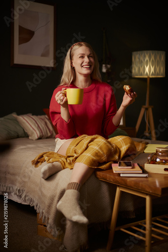 Cuadro en lienzo Vertical image of happy Caucasian young woman sitting on bed, holding mug and eating snack