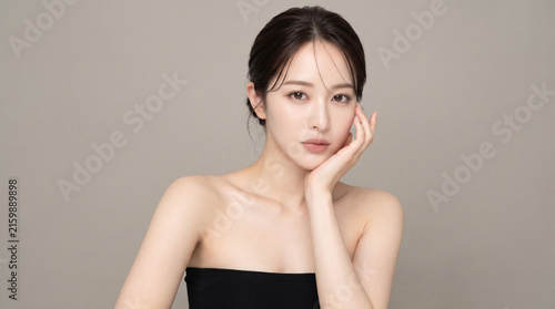 トープカラーの背景で頬に手を添えながら凛とした表情を見せる若い日本人女性、黒いベアトップと透明感のある素肌を生かした美容・コスメ広告向けポートレート