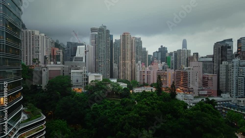 Drone footage overlooking densely packed high-rise residential buildings in Hong Kong, with a sports complex, urban roads, and green mountains in the background.